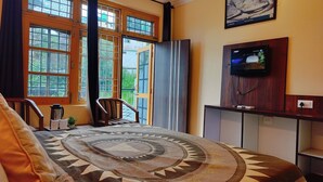 A room with a large window, a bed with a patterned rug, a wooden chair, a coffee maker, and a TV on a wooden stand. - Ishwar Guest House (Dharamshala)
