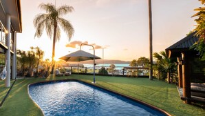 A swimming pool with a view of the sunset over a body of water, surrounded by palm trees and a grassy area with outdoor furniture.