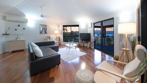 A spacious living room with a black sofa, a round coffee table, and a large TV. There is a sliding glass door leading to a balcony with a view of the outdoors.