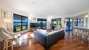 A spacious living room with a large window offering a view of the sea, a modern sofa, a dining area with a bar, and a television.