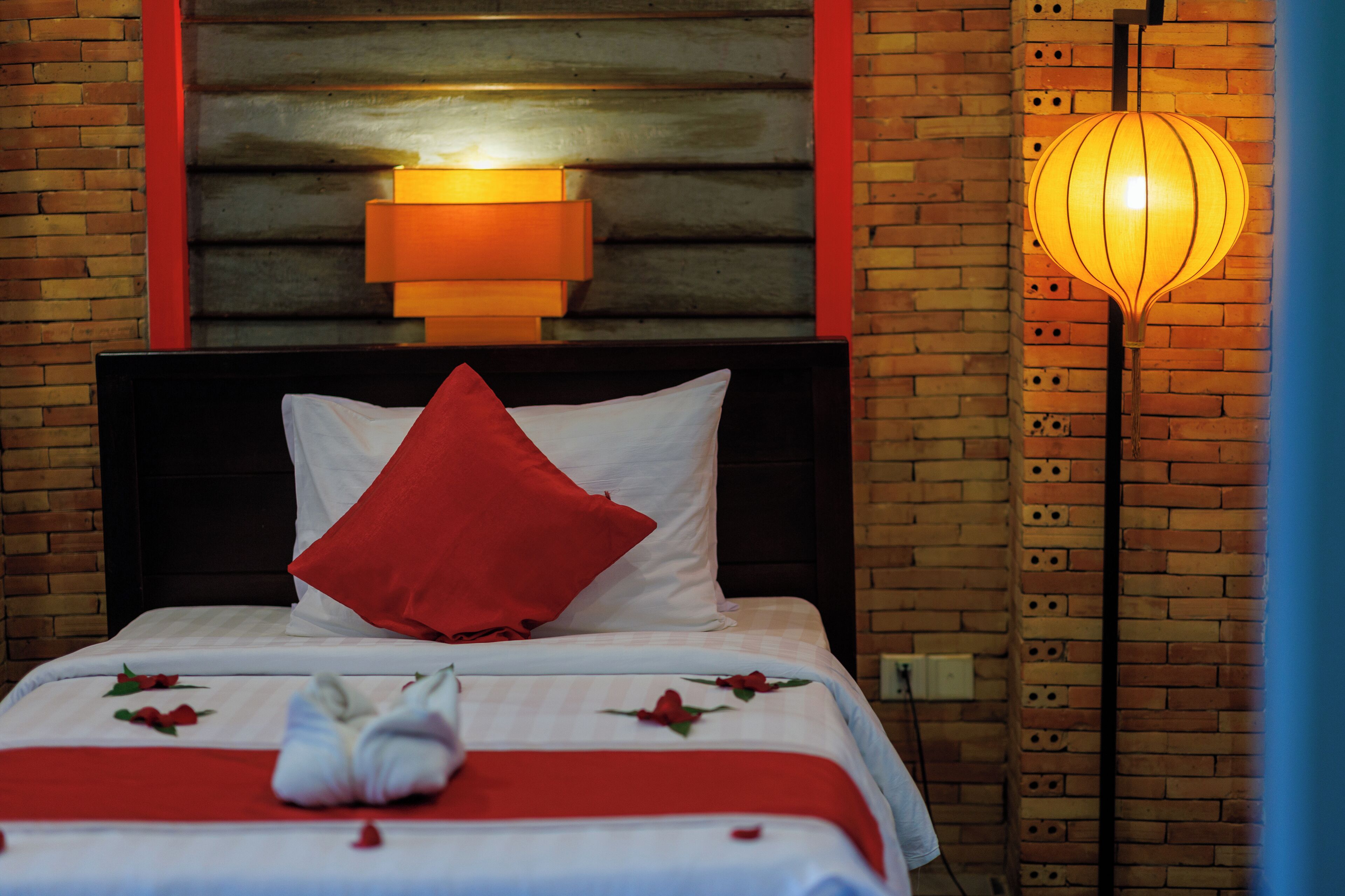 A neatly made bed with a red pillow, a white pillow, and a red and white bedspread with rose petals. There is a wall-mounted light fixture above the bed and a floor lamp to the right.
