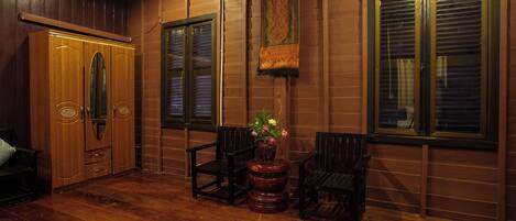 A room with wooden panel walls, a wooden wardrobe, two chairs, a small table with a vase of flowers, and a window with blinds.
