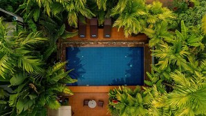 An aerial view of a rectangular swimming pool surrounded by lush tropical plants, with a wooden deck and two small tables. - RESIDENCE 1960 (Siem Reap)