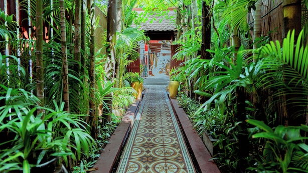 A narrow pathway lined with lush greenery and a patterned tile floor leading to a traditional structure. - RESIDENCE 1960 (Siem Reap)