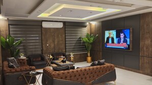 A modern living room with a large sectional sofa, a coffee table, and a television displaying a news broadcast. - Royal Group Hotel Irbid (Irbid)