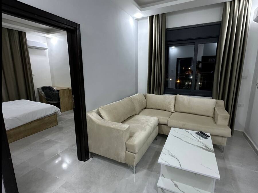 Una sala de estar moderna con un sofá seccional color beige, una mesa de centro de mármol blanco y una cama con cabecera de madera.