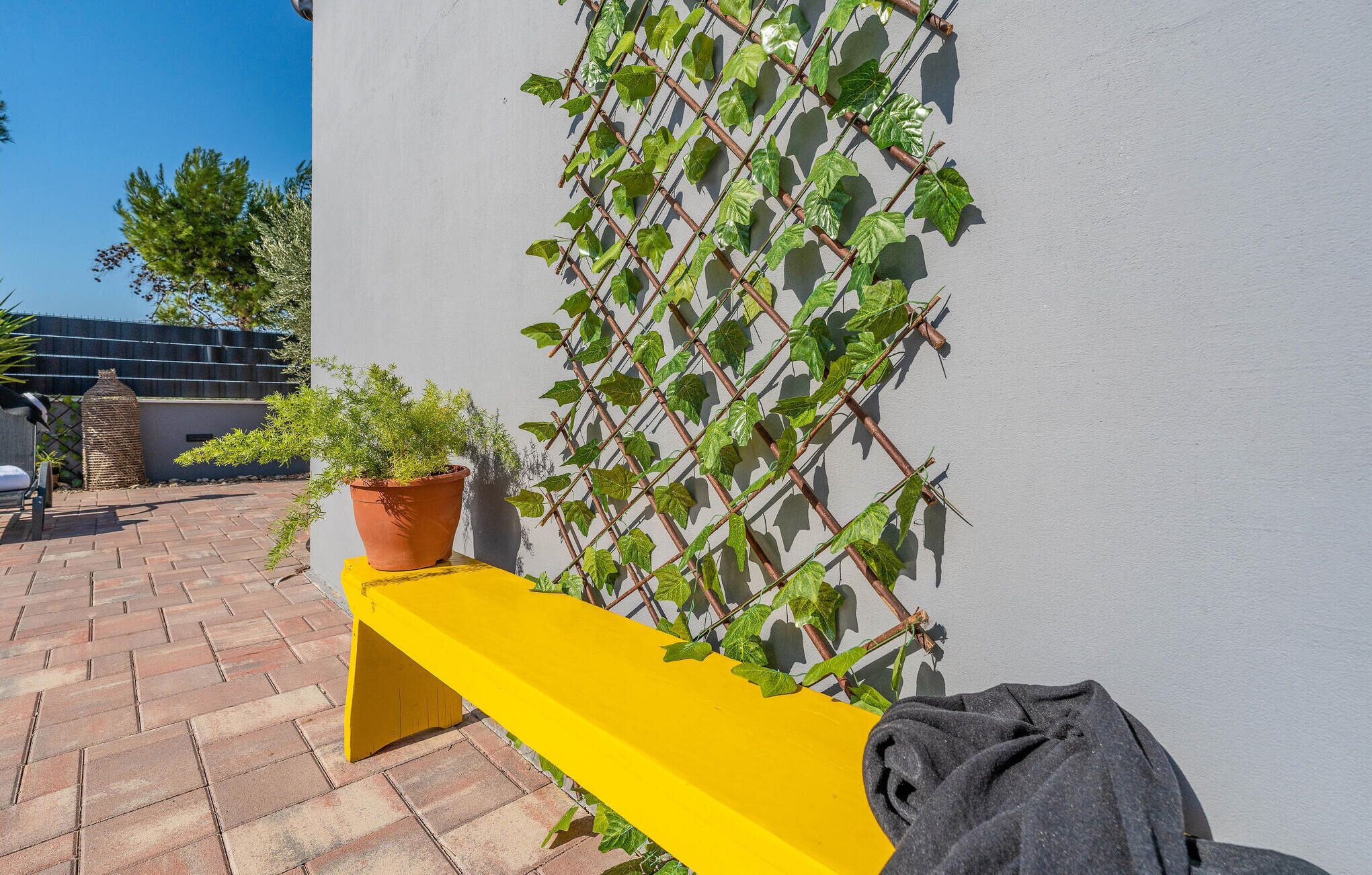 Un banc jaune, avec un pot de fleurs à côté, une plante grimpante sur un mur et une terrasse carrelée.