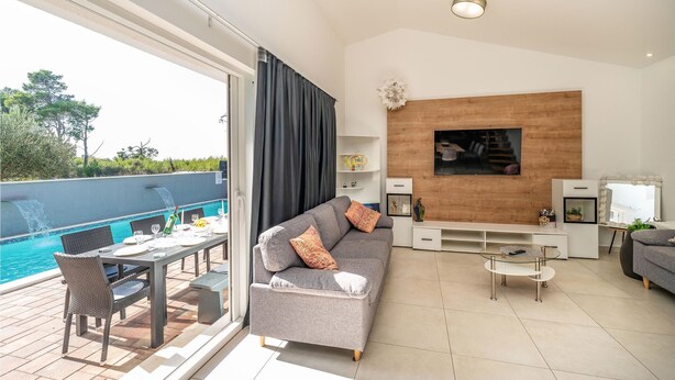 Un salon moderne avec un canapé gris, une table basse en verre et un téléviseur fixé sur un mur en bois. On peut apercevoir une piscine à travers la porte vitrée.