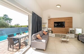 A modern living room with a grey sofa, a glass coffee table, and a TV mounted on a wooden wall. There is a pool visible through the glass door. - Beautiful home in Privlaka (Privlaka)
