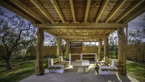 Un pavillon en bois installé à l’extérieur, doté de bancs et d’une table, et entouré d’arbres et d’une clôture.