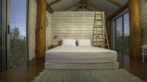 Une chambre avec un grand lit, des poutres en bois, une échelle et une vue dégagée sur l’extérieur grâce à de grandes fenêtres.