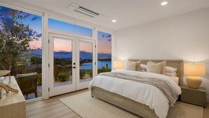 A bedroom with a large bed, two bedside lamps, and a view of a lake and mountains through glass doors.