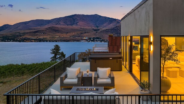 A modern outdoor deck with wicker furniture, a fire pit, and a view of a lake and mountains.