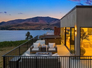 Une terrasse extérieure moderne, dotée de meubles en osier, d’un foyer et offrant une vue sur un lac et des montagnes.