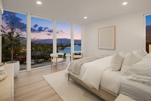 A bedroom with a large window, a bed, a chair, and a plant.