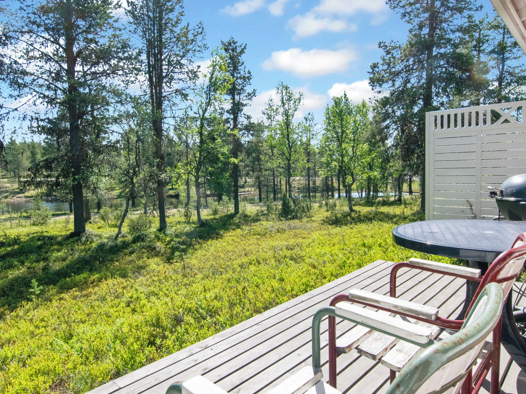 Une terrasse en bois avec une table et des chaises, donnant sur une zone verdoyante avec des arbres et une étendue d’eau.