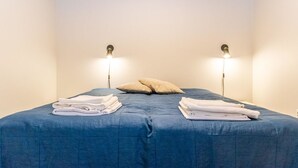 Un lit bien fait, avec une literie bleue et deux oreillers, de part et d’autre duquel se trouvent deux lampes fixées au mur.