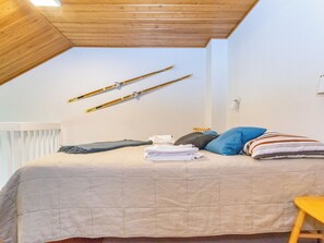 Une chambre avec un plafond en pente, un lit avec des oreillers et des vêtements pliés, et des paires de skis appuyés contre le mur.