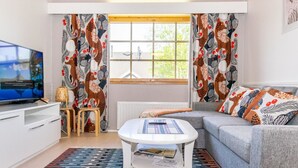 Un salon moderne doté d’un téléviseur à écran plat, d’une table basse blanche, d’un canapé gris, de rideaux à motifs et d’une fenêtre donnant sur un paysage verdoyant.