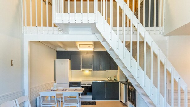 Une cuisine moderne avec des meubles sombres, un réfrigérateur blanc et une cuisinière à gaz. On y trouve un coin repas avec des chaises et une table blanches, ainsi qu’un escalier avec des rampes blanches menant à l’étage supérieur.