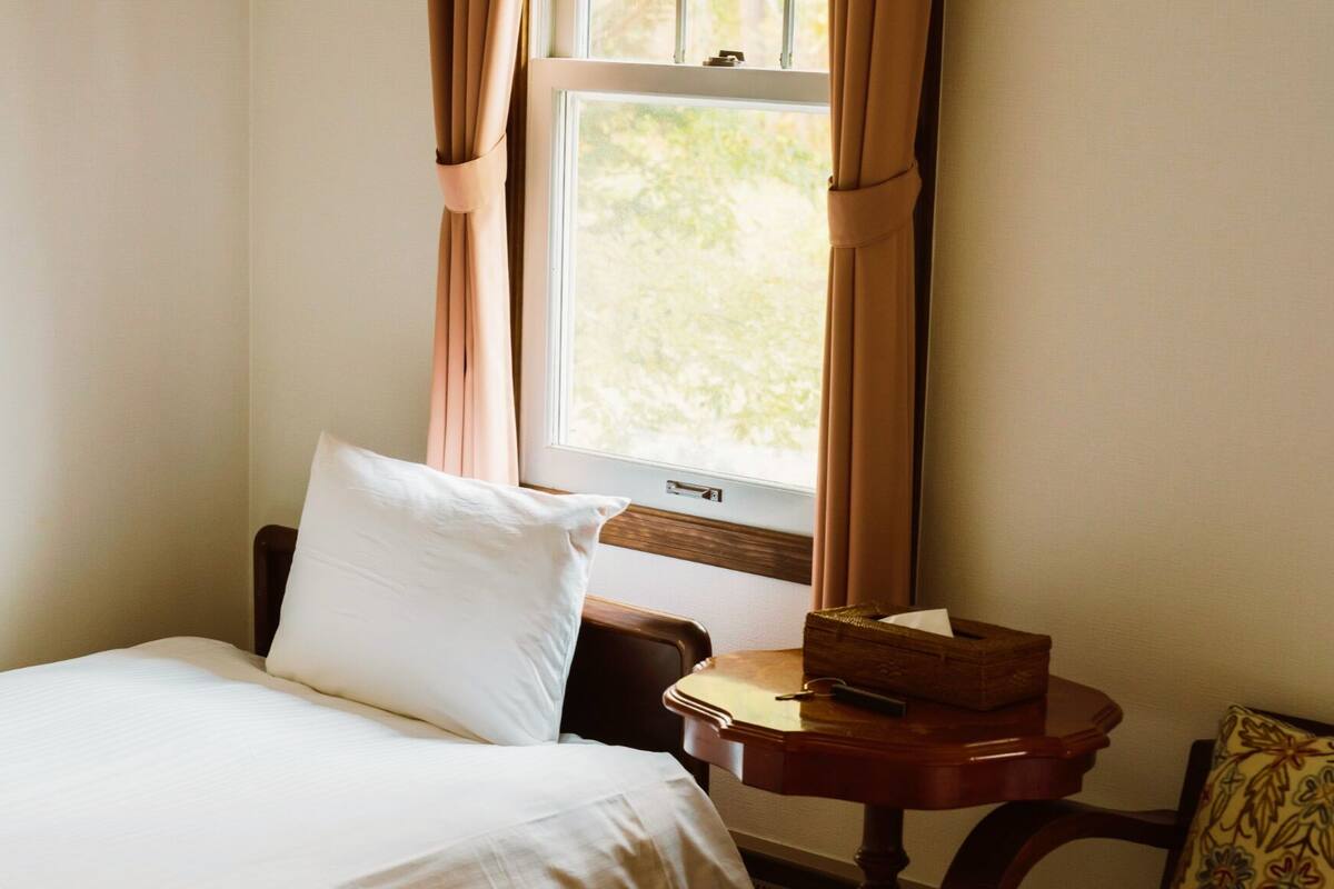 A hotel room with a bed, a bedside table with a lamp, a window with curtains, and a view of greenery outside.