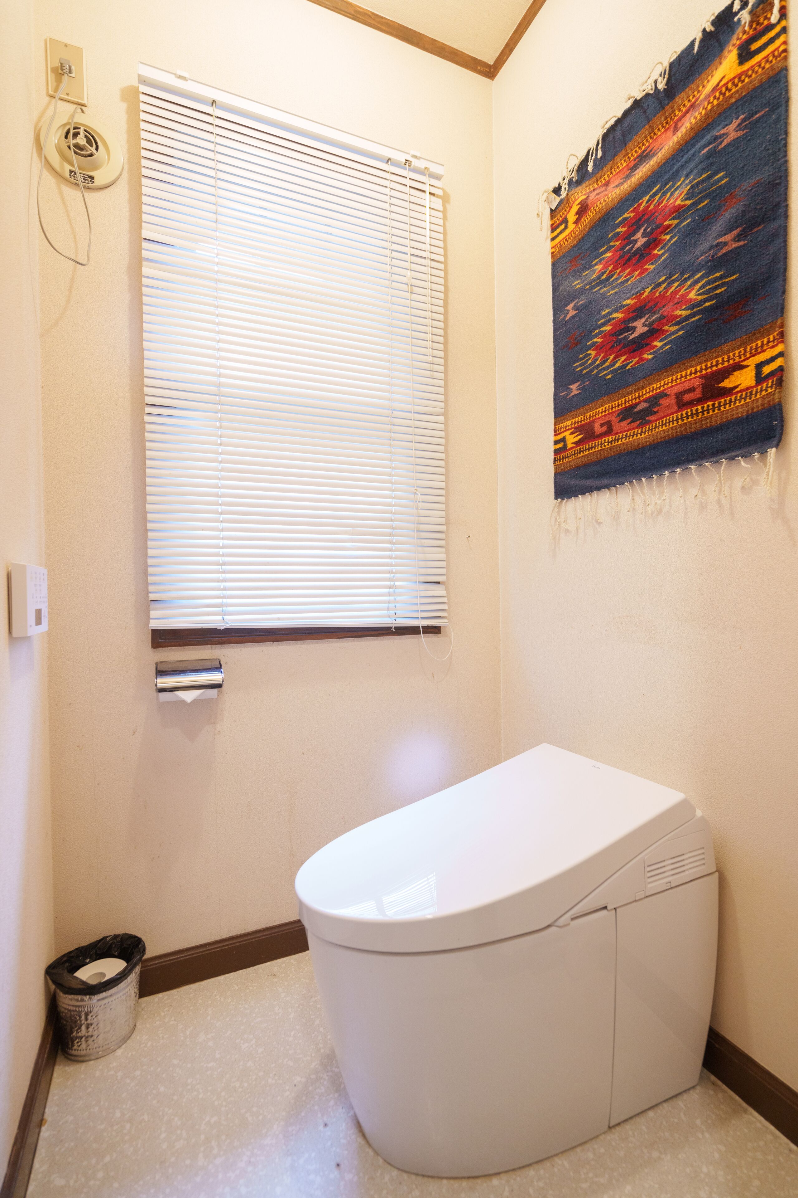 A bathroom with a toilet, a window with blinds, a wall-mounted heater, and a decorative tapestry.