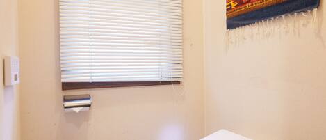 A bathroom with a toilet, a window with blinds, a wall-mounted heater, and a decorative tapestry.