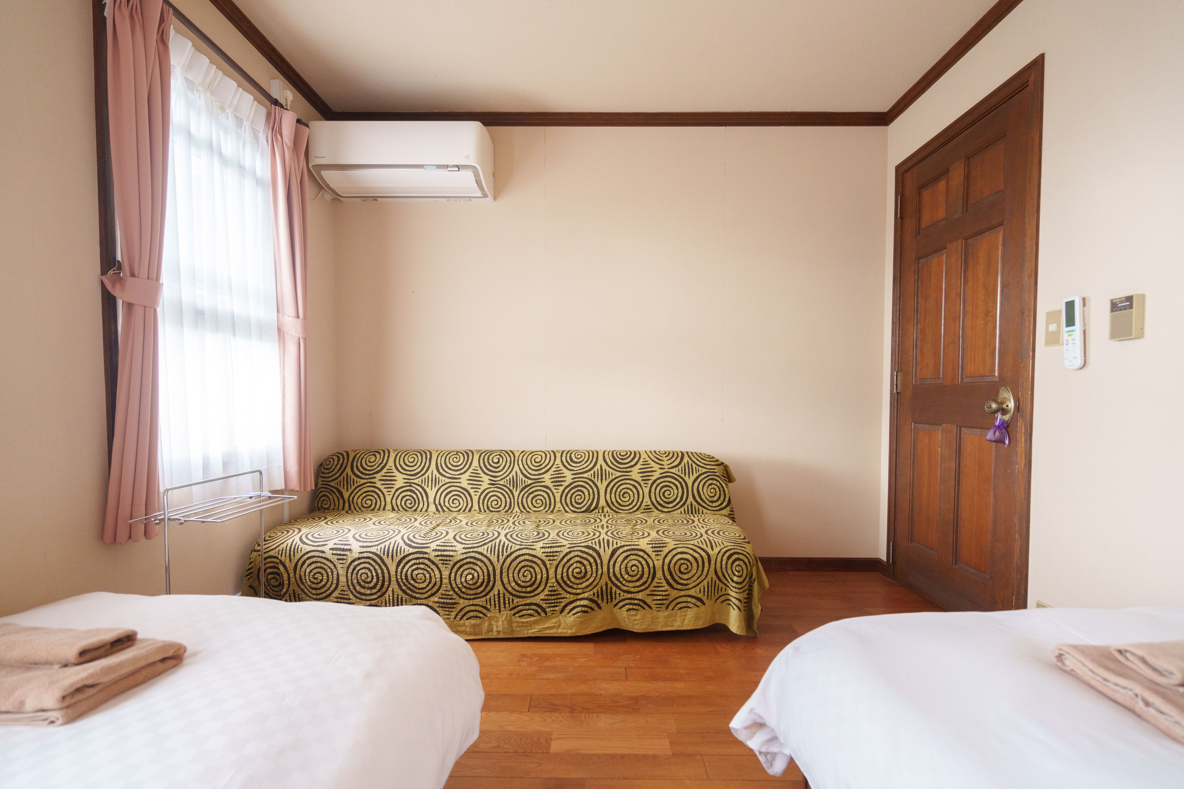 A hotel room with two beds, a sofa, an air conditioning unit, a wooden door, and a window with curtains.