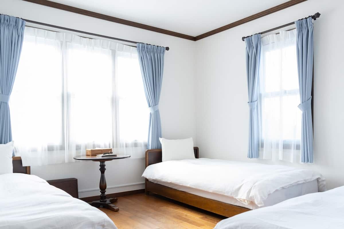 A hotel room with three single beds, a small table, and two windows with curtains.