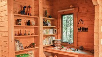 A cozy cabin interior with wooden shelves filled with books, puzzles, and toys. A window with a view of trees is visible.