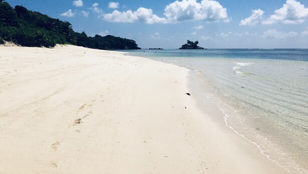 Una playa extensa y arenosa con agua turquesa cristalina, una isla a lo lejos y una zona boscosa.
