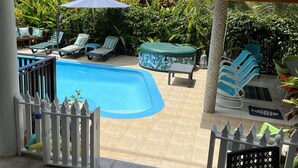 Un área de piscina con sillones de descanso y sombrillas, rodeada de exuberante vegetación y montañas al fondo.