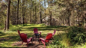 A forested area with a small table and two Adirondack chairs.