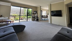 A spacious living room with grey sofas, a flat-screen TV mounted on the wall, and a large sliding glass door leading to a patio area with outdoor furniture. - Comfortable family home, Privacy & convenience, pets welcome! (Papamoa)
