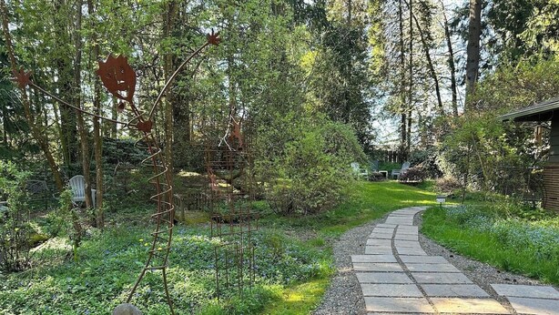 A garden pathway leading to a rustic cabin, surrounded by dense greenery and a metal sculpture.