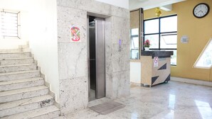 A marble staircase leading to an elevator, a reception desk with a no smoking sign, and a clock on the wall. - Smile stay Karanji Comforts (Bengaluru)