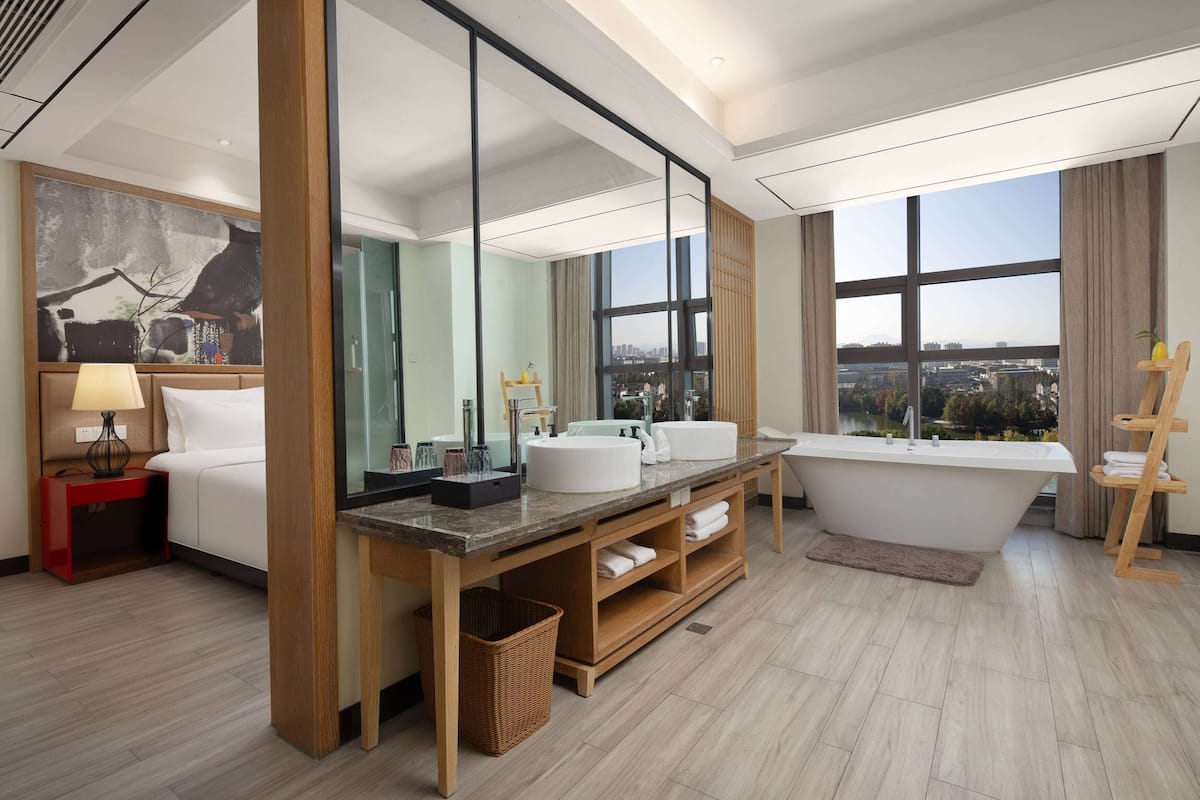 A modern hotel bathroom with a large bathtub, dual sinks, a large mirror, and a view of the city.