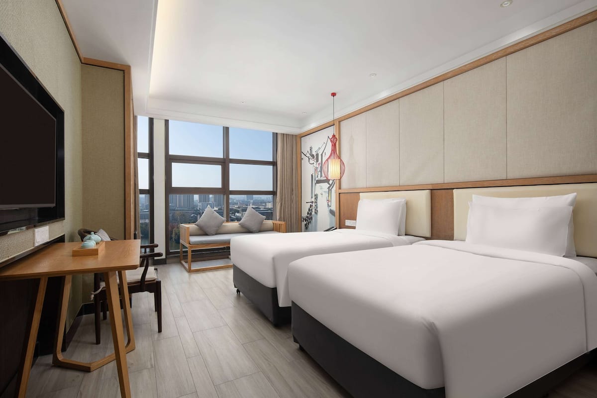 A hotel room with two beds, a desk, and a large window offering a city view.