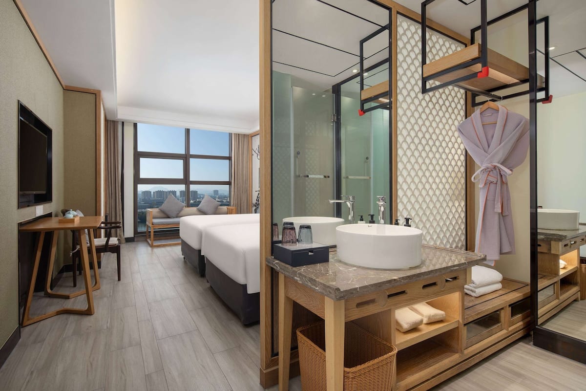 A modern hotel room with a large bed, a sitting area, a bathroom with a large mirror, and a view of the city.