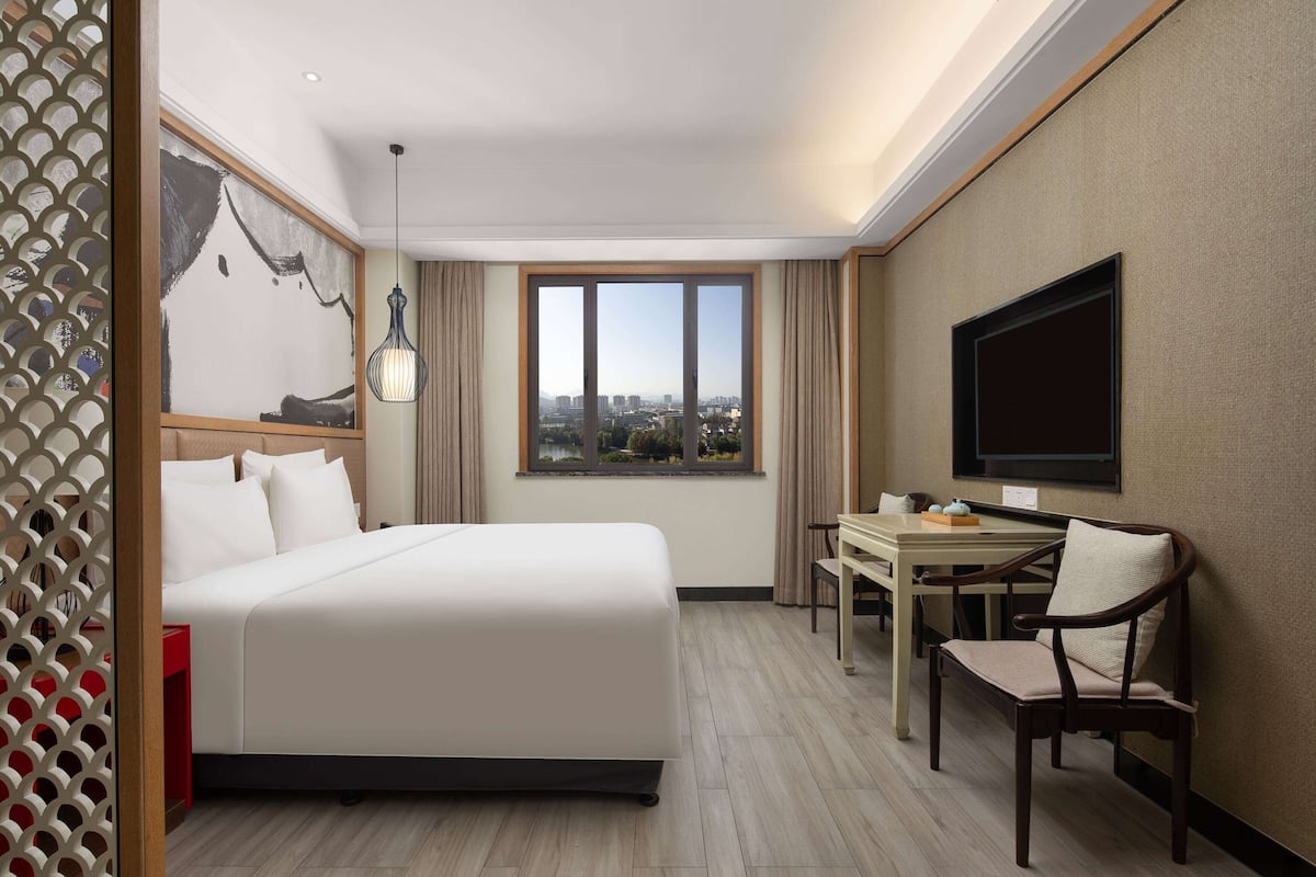 A hotel room with a bed, a desk, a chair, a TV, and a window with a city view.