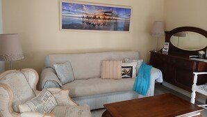 Una sala de estar con un sofá color beige, una mesa de centro de madera y un sillón antiguo. En la pared, un cuadro de un atardecer sobre un cuerpo de agua.