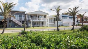 A modern two-story building with a balcony, surrounded by lush greenery and palm trees. - On The Beach (Durban North)