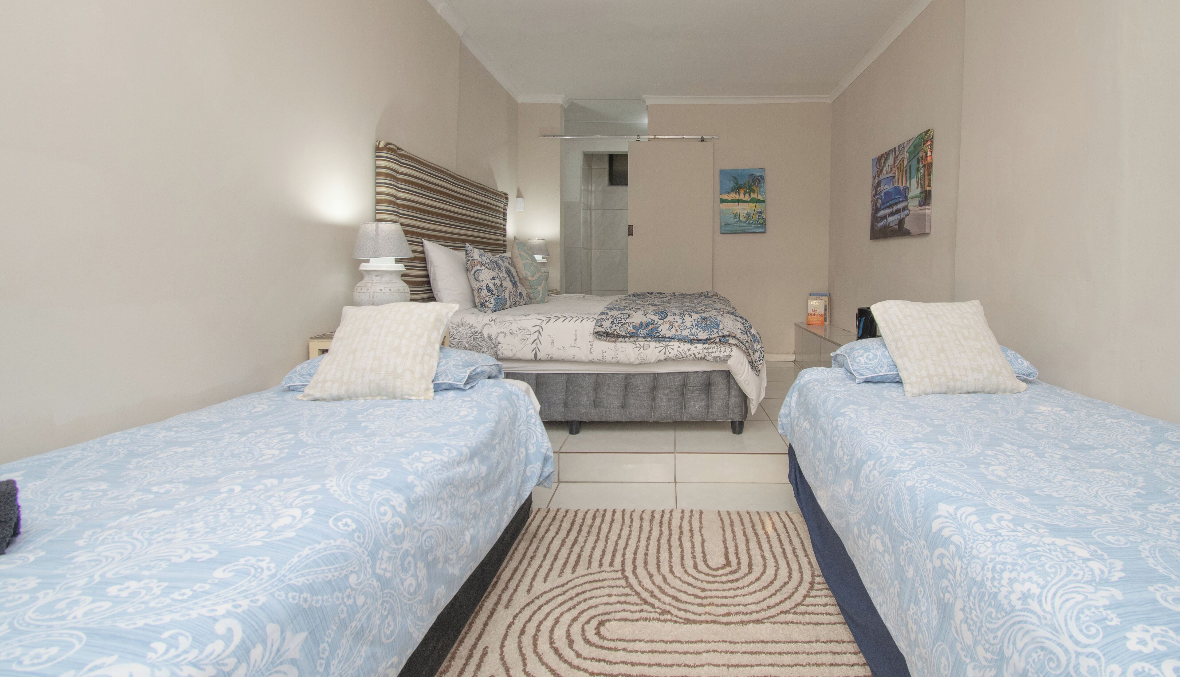 A bedroom with two single beds, a nightstand, a lamp, a small table, and a door leading to another room.