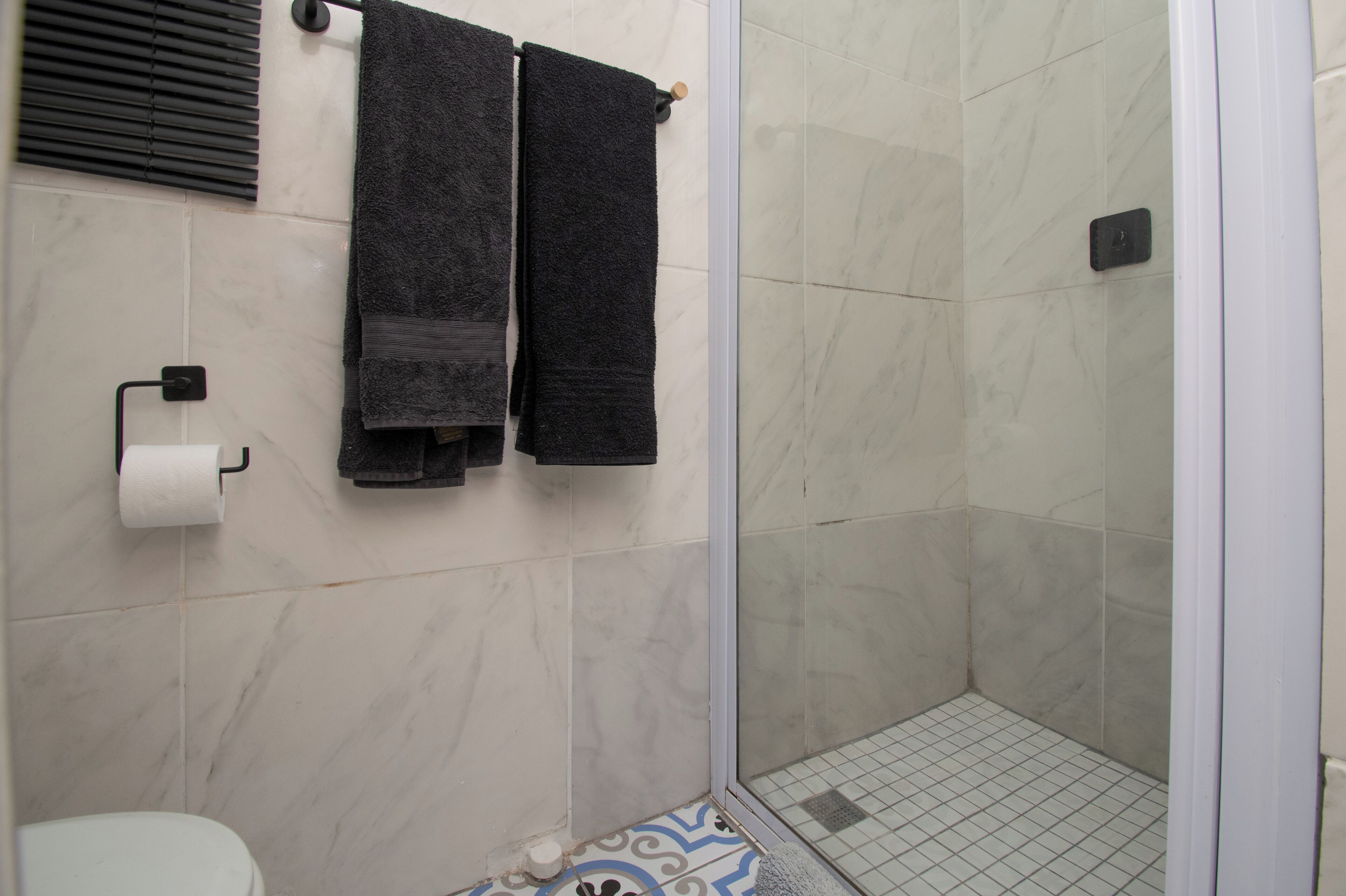 Une salle de bain avec des toilettes, deux serviettes suspendues à un porte-serviettes, un espace douche doté d’une porte en verre et un carrelage décoratif.