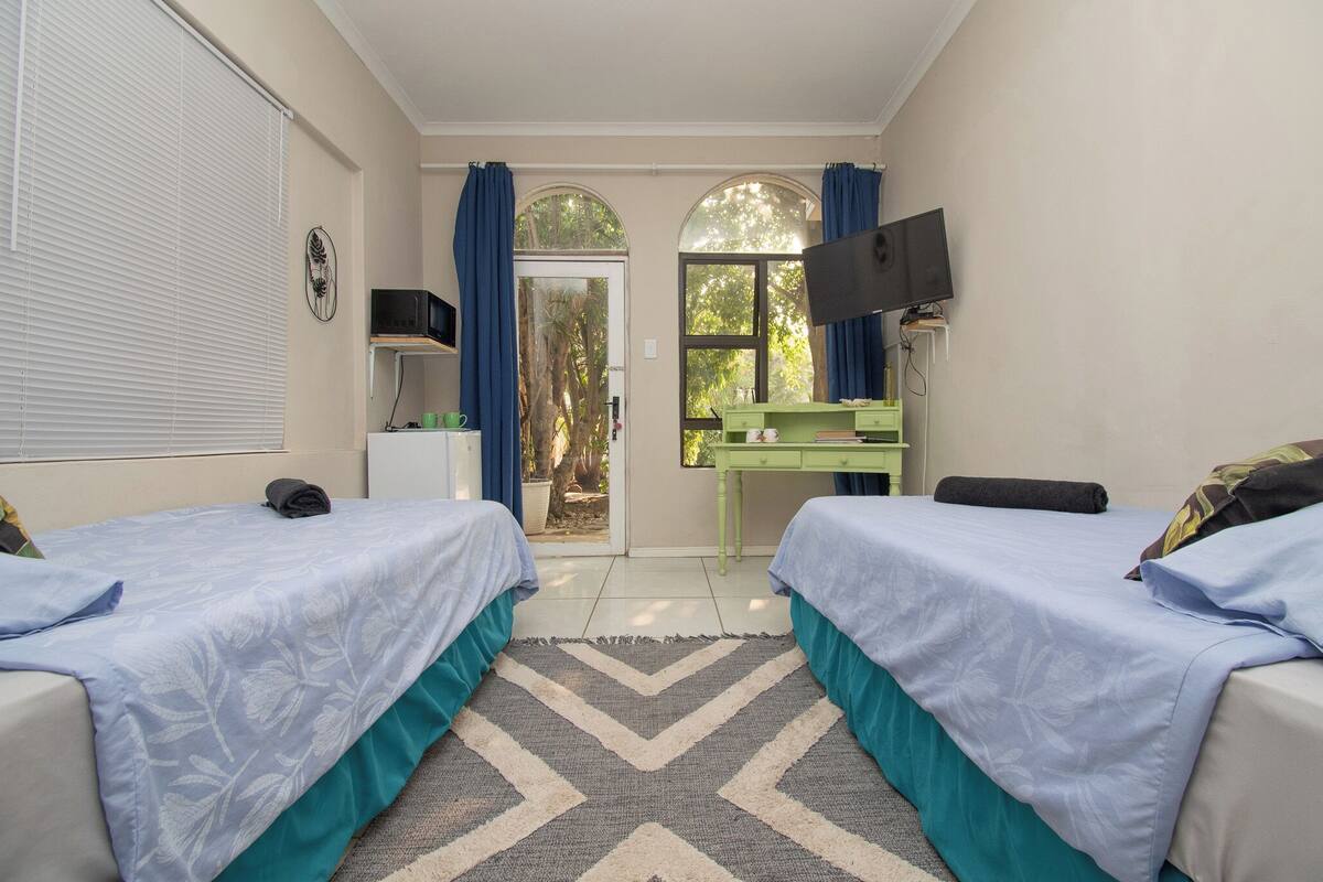 Une chambre avec deux lits, un bureau, une télévision et une porte donnant sur un espace extérieur.
