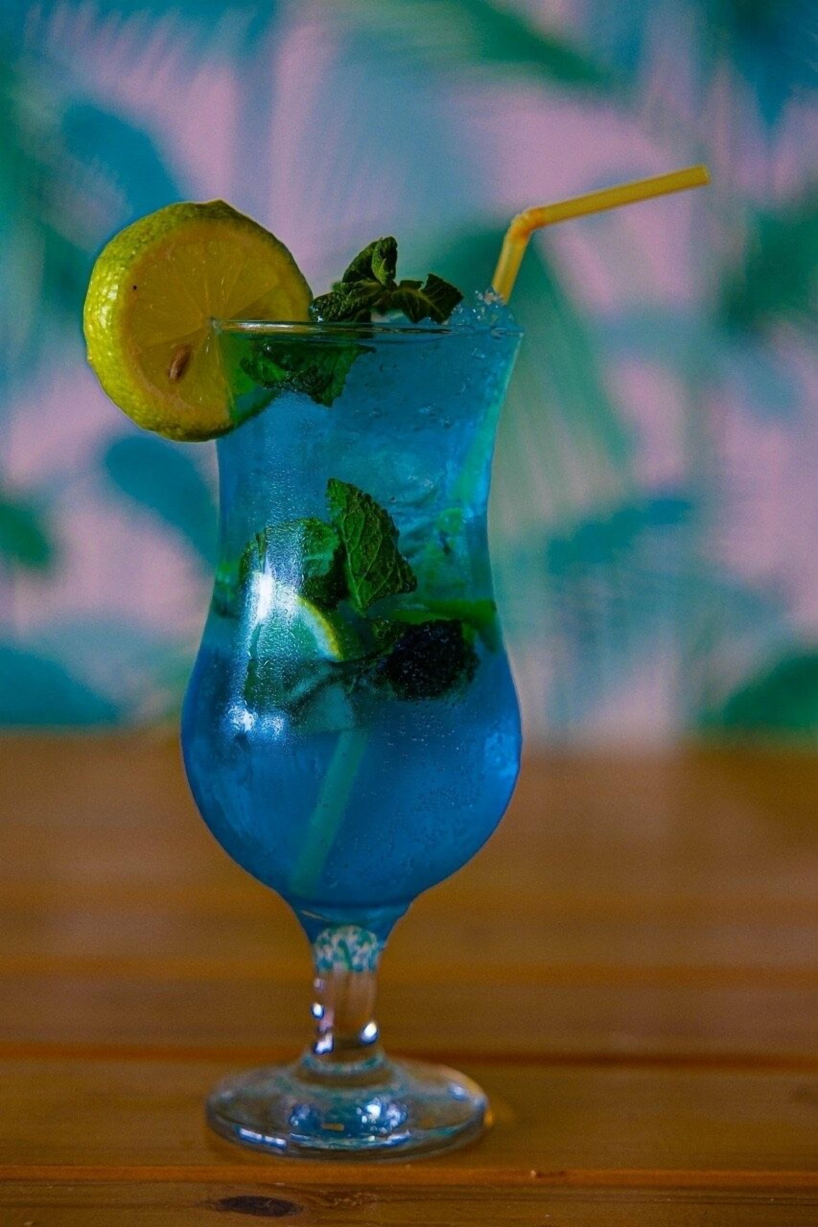 A tall glass with a blue drink, garnished with a lemon slice and mint leaves, with a straw.