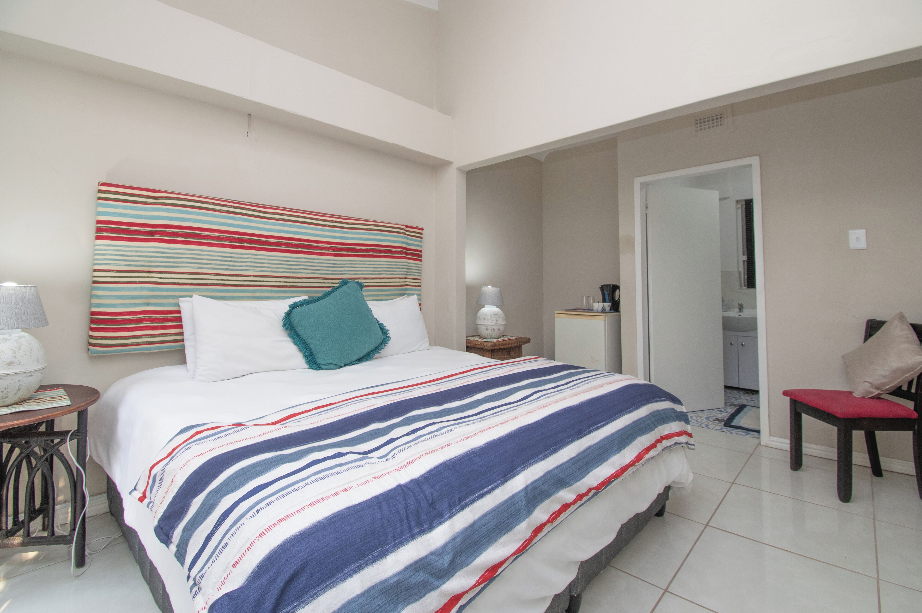 A bedroom with a large bed, a bedside table with a lamp, a chair, and a bathroom visible through an open door.