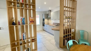 A modern kitchen with white cabinets, a central island, and a dining area with a wooden table and two turquoise chairs. - Modern House Near Beach: 4 Bedrooms, Full Kitchen, WiFi, Terrace, Parking (Bretignolles-sur-Mer)