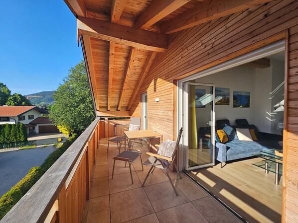 A wooden balcony with a table and chairs, a sliding glass door leading to a living area with a sofa and cushions, and a view of houses and greenery.