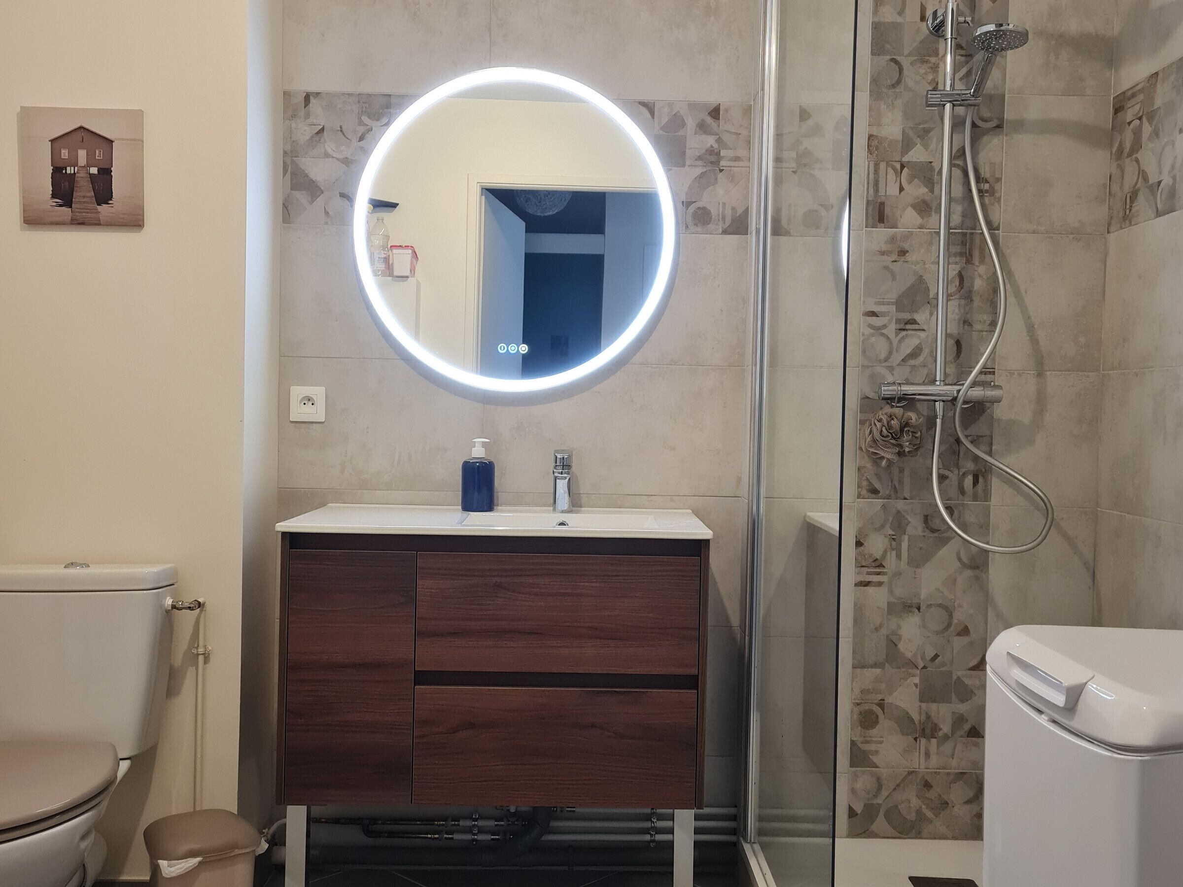 Une salle de bain avec un miroir rond, des toilettes, un lavabo avec un distributeur de savon et une douche.